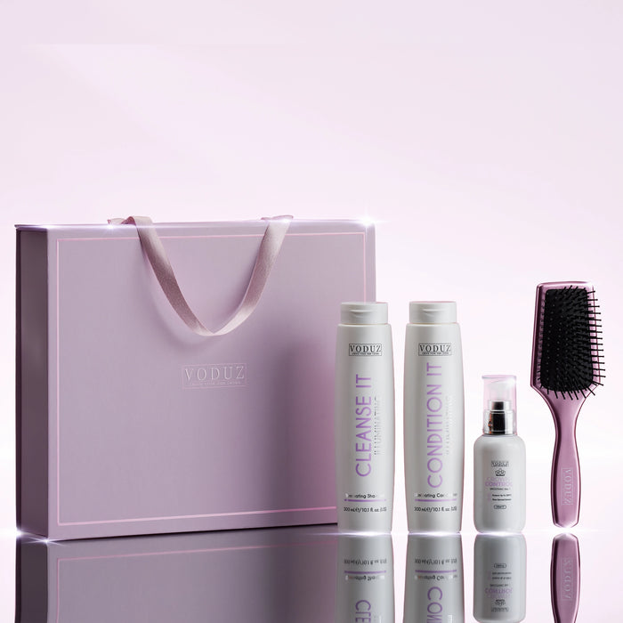 VODUZ hair care gift set featuring a lavender gift bag, two white bottles labeled Cleanse It and Condition It, a small white spray bottle, and a lavender hairbrush, arranged on a reflective surface against a soft lavender background.