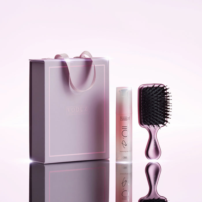 A VODUZ  hair care gift set featuring a translucent lavender hairbrush, a matching hair oil bottle, and a lavender gift box with ribbon handles, displayed on a reflective surface against a light pink background.