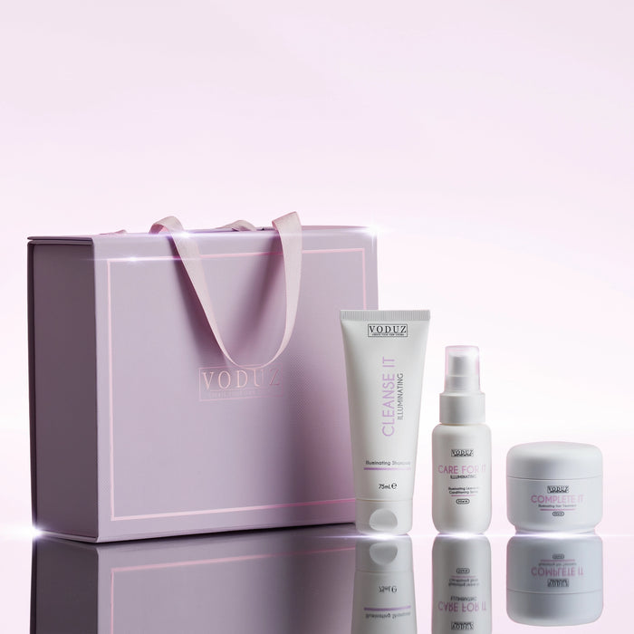 VODUZ skincare gift set featuring a lavender gift bag with pink handles and three white skincare containers (Cleanse It tube, Care For It spray, and Complete It cream) arranged on a reflective surface against a soft pink background.