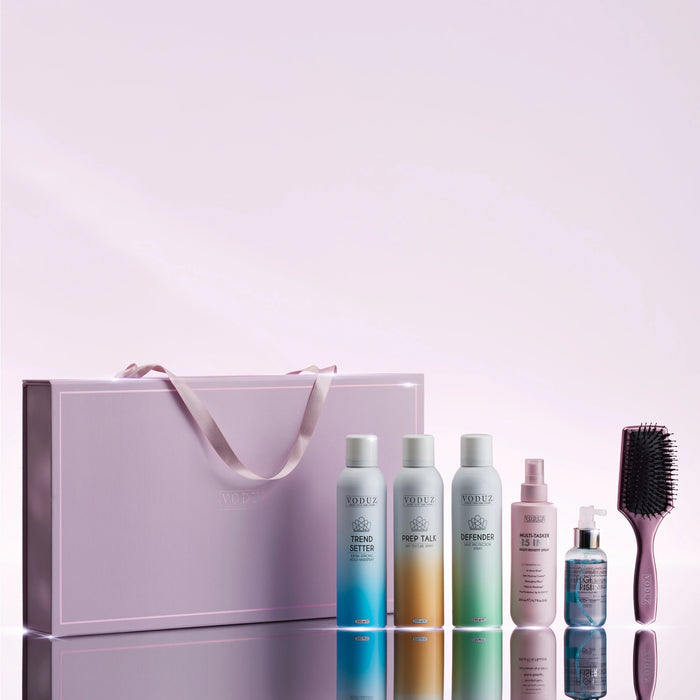 A haircare gift set from VODUZ Haircare arranged on a reflective surface against a soft pink studio background. The set includes a large lavender gift bag with a ribbon, three gradient bottles labeled 'TREND SETTER', 'PREP TALK', and 'DEFENDER,', a pink spray bottle of 25-IN-1 Multi-Tasker, a small blue bottle of HIGH RISER, and a lavender hairbrush.